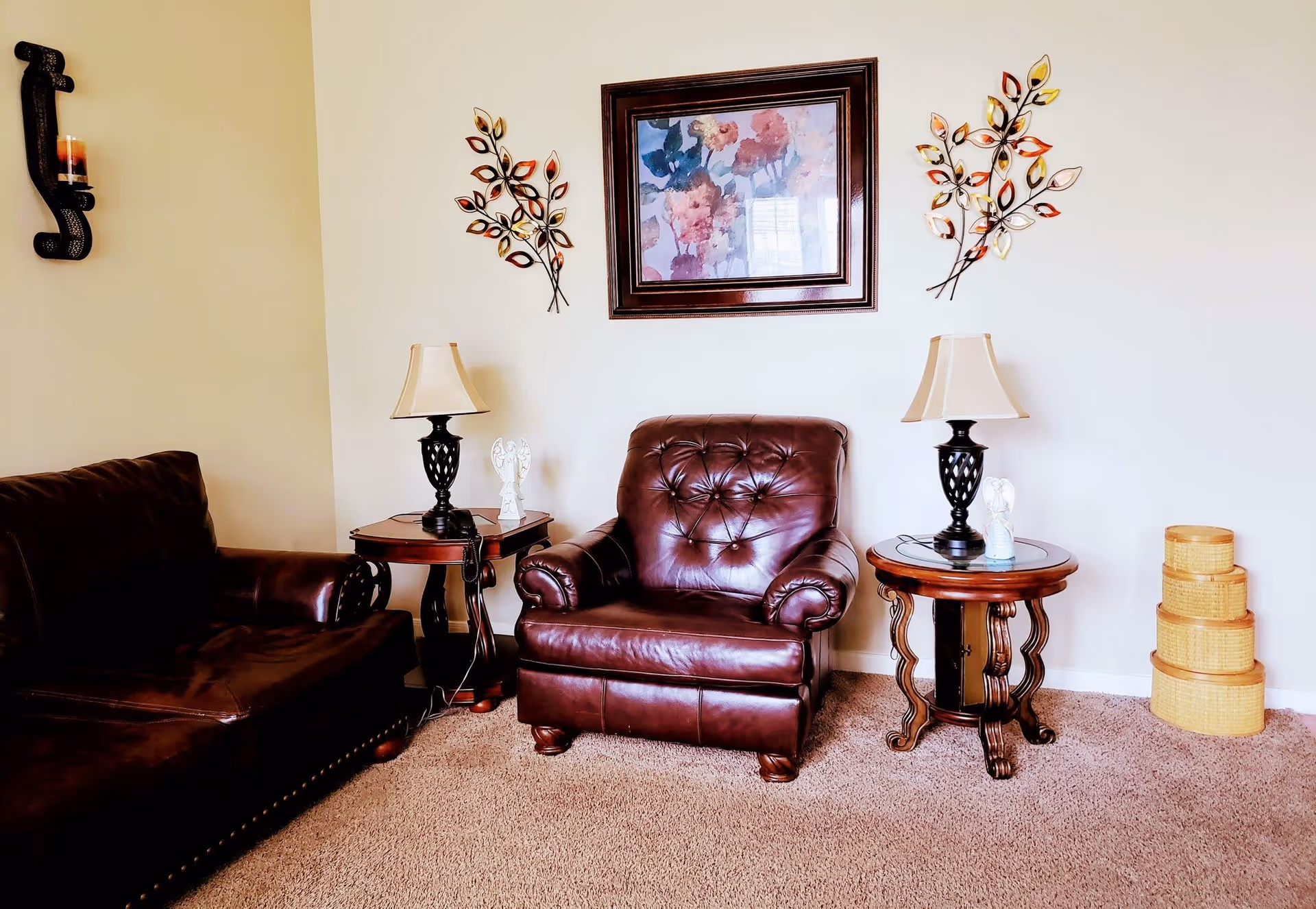 A cozy living room area with a dark brown leather armchair and a matching sofa. Two wooden side tables with lamps and decorative angel figurines flank the armchair. Above the armchair is a framed floral painting, flanked by two metal wall decorations shaped like branches with colorful leaves. On the right side of the image, there are three stacked woven baskets on the carpeted floor.
