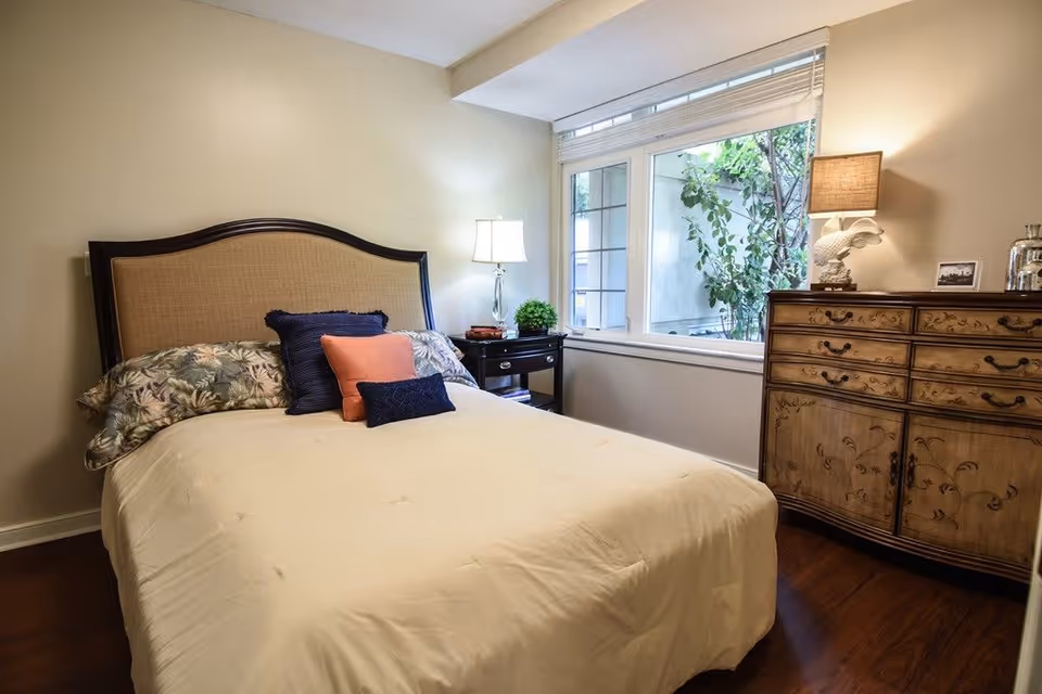A cozy bedroom with a large bed featuring a beige comforter and multiple pillows in floral, navy blue, and coral colors. There is a wooden nightstand with a lamp and a small plant next to the bed. A large window with white blinds lets in natural light, and a wooden dresser with decorative carvings and a lamp sits against the wall.