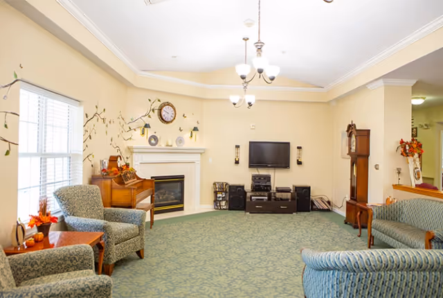 A cozy senior living common area with patterned armchairs and a sofa arranged around a carpeted floor. There is a fireplace with a clock and decorative plates above it, a wall-mounted TV with speakers and media equipment below, and a wooden grandfather clock. The room is softly lit with ceiling lights and has a window with blinds letting in natural light. Autumn-themed decorations are placed on tables and surfaces.