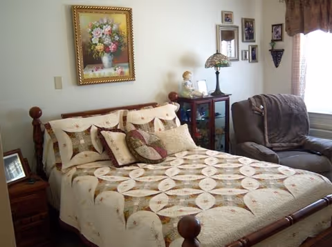 A cozy bedroom featuring a wooden bed with a patterned quilt and multiple pillows. Next to the bed is a wooden nightstand with a framed photo. On the wall above the bed hangs a framed floral painting. A glass-front cabinet with decorative items and a Tiffany-style lamp sits beside a comfortable armchair draped with a blanket. Several framed pictures and a small shelf adorn the adjacent wall near a window with curtains.