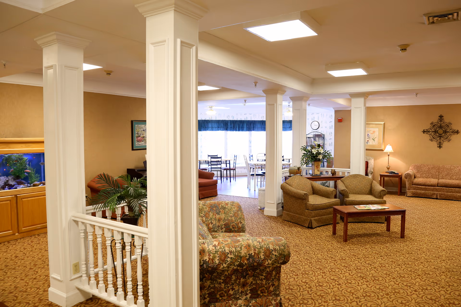 A spacious and well-lit common area in a senior living facility featuring multiple seating arrangements with armchairs and sofas, a wooden coffee table with magazines, decorative plants, a large fish tank, and a dining area with tables and chairs in the background near large windows.