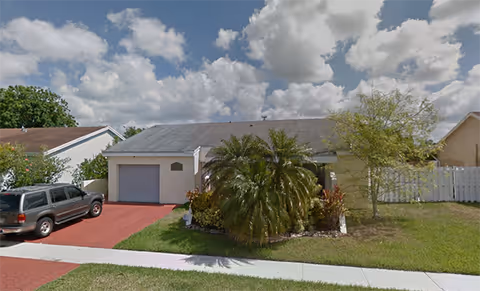 Single-story residential building with a gray roof, a garage on the left, a driveway with a parked SUV, and a front yard featuring a large palm tree and other greenery under a partly cloudy sky.