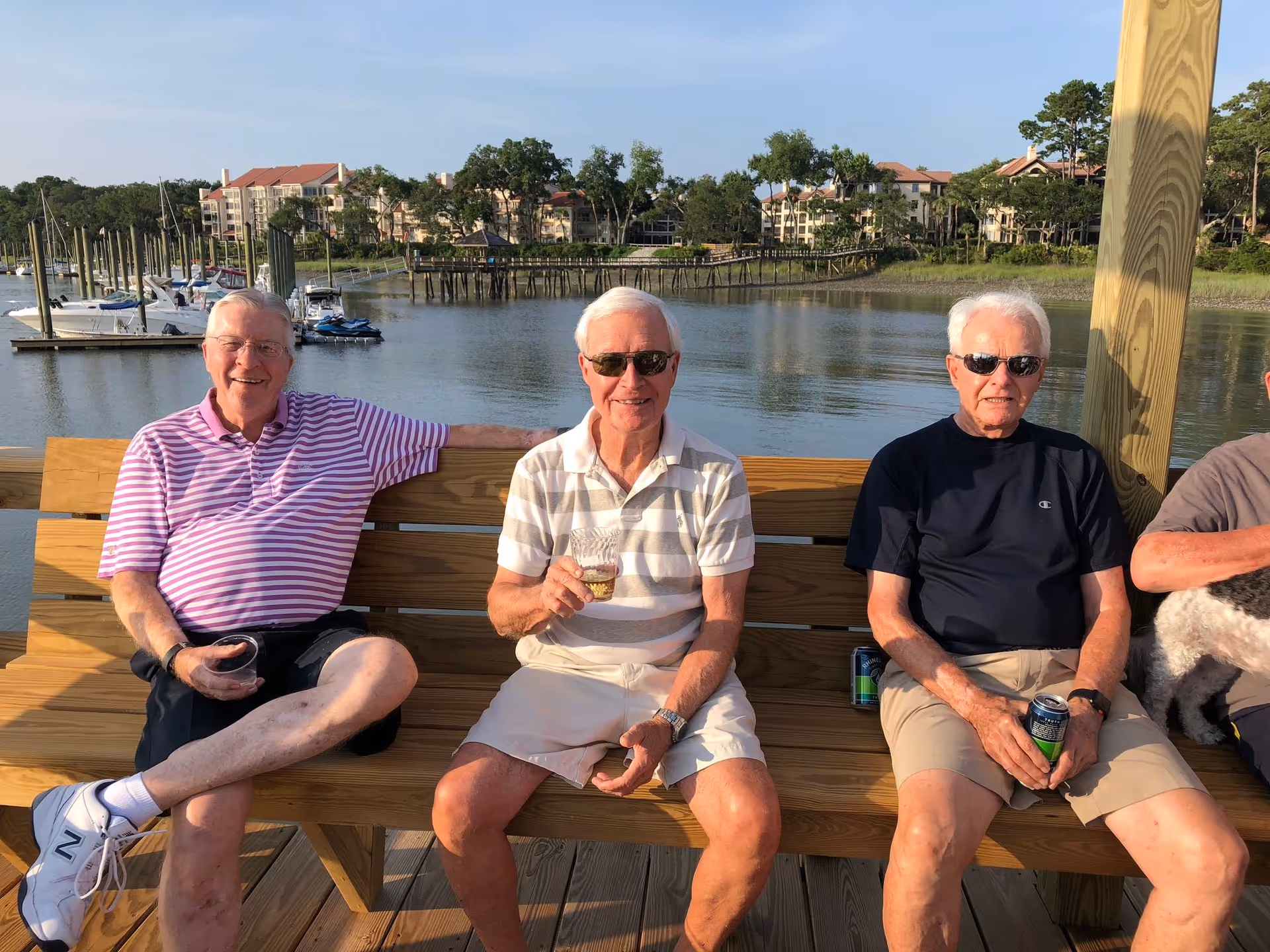 Three elderly men sitting on a wooden bench on a dock by the water, with boats and buildings in the background. They are enjoying drinks and wearing casual summer clothing and sunglasses.