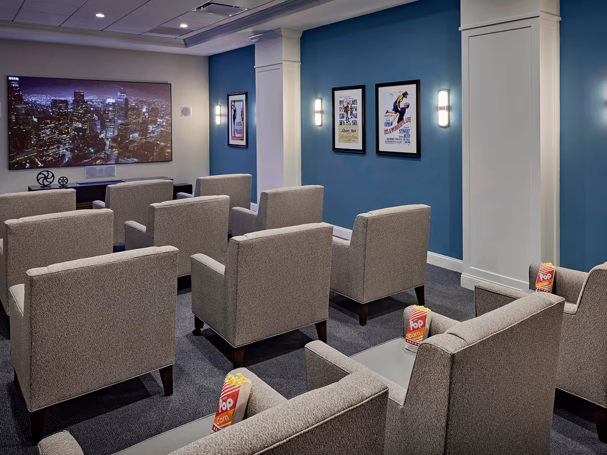 A small movie theater room with rows of gray upholstered armchairs facing a large screen displaying a cityscape at night. The walls are painted blue and white, with framed movie posters and wall sconces providing lighting. Some chairs have popcorn bags placed on the armrests.