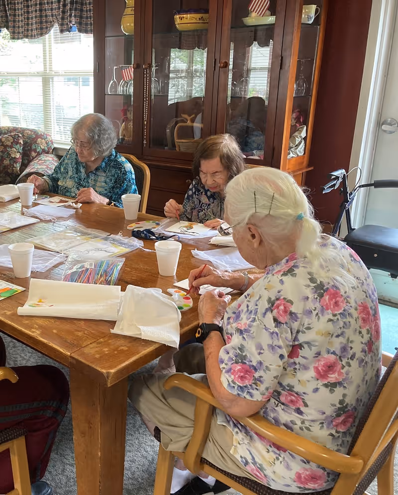 Three elderly women sitting around a wooden table engaged in a painting activity. The table has art supplies including paint palettes, brushes, and paper towels. Behind them is a wooden cabinet with glass doors displaying dishes and glassware. A window with checkered curtains lets in natural light.