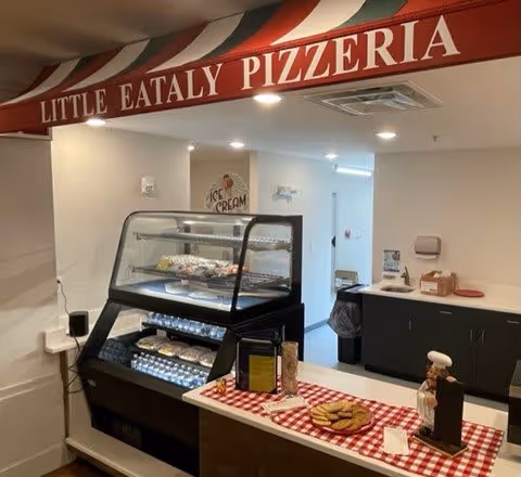Interior view of a small pizzeria area named Little Eataly Pizzeria inside a senior living facility. The area features a display case with bottled water and food items, a counter with a red and white checkered cloth holding cookies and a small chef figurine, and a sign on the wall that says Ice Cream.