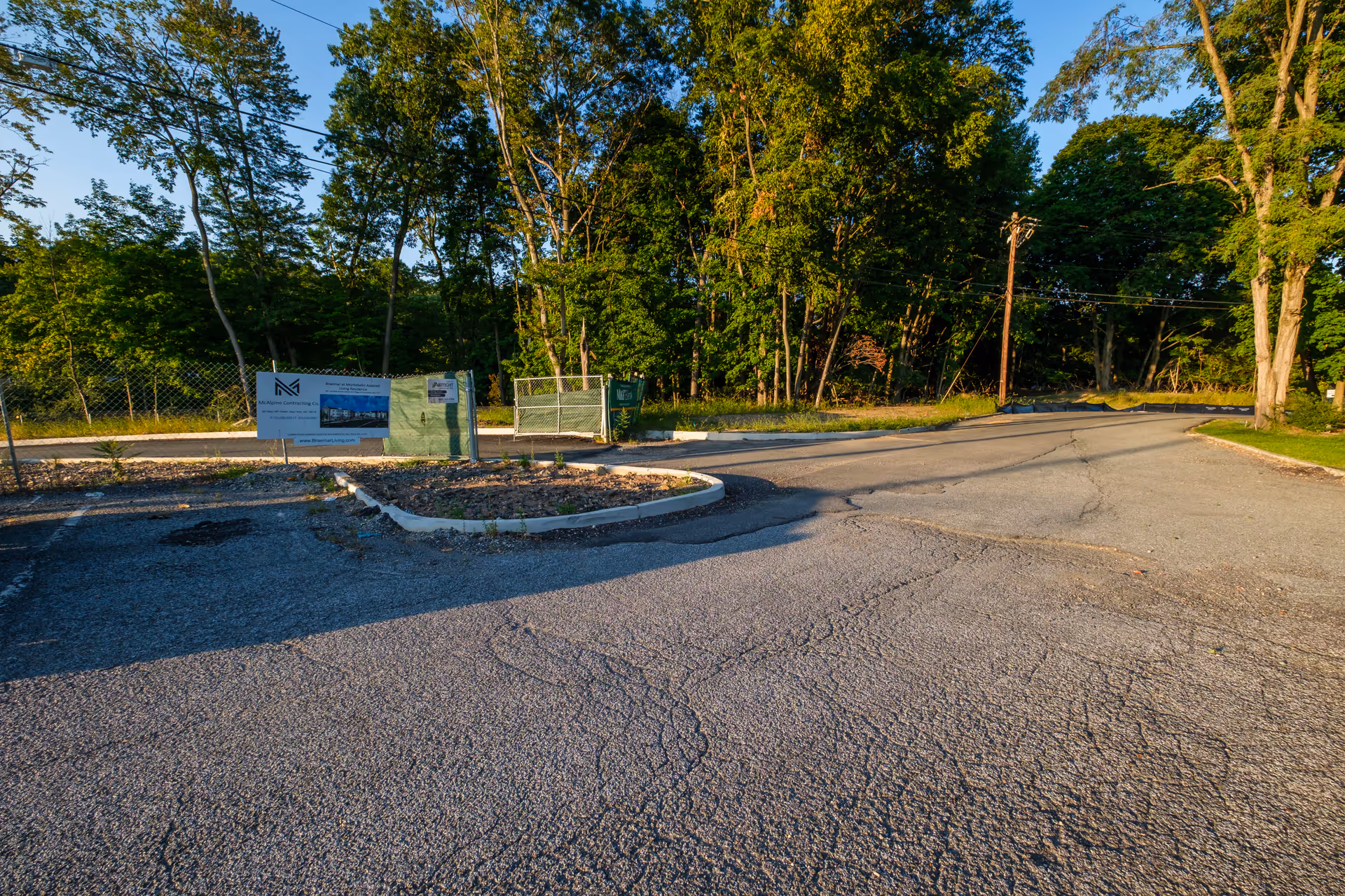 An outdoor paved road leading to a fenced area with construction signs, surrounded by tall green trees under a clear blue sky.