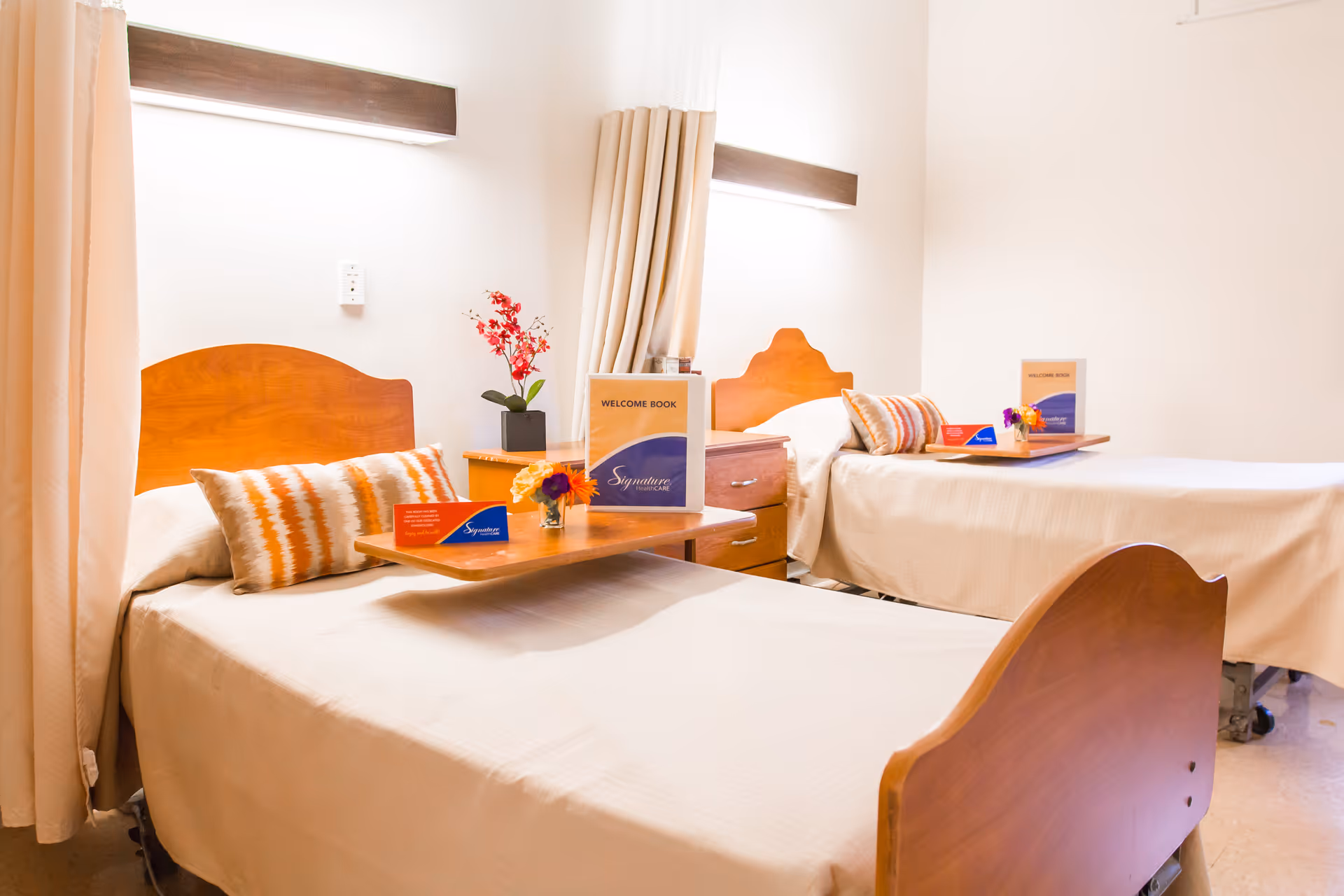 A bright and clean nursing facility room with two single beds separated by a wooden nightstand. Each bed has a beige bedspread, a decorative pillow, and a wooden headboard. Overhead lights illuminate the beds, and there are small flower arrangements and welcome books placed on wooden overbed tables. Light-colored curtains hang beside the beds.