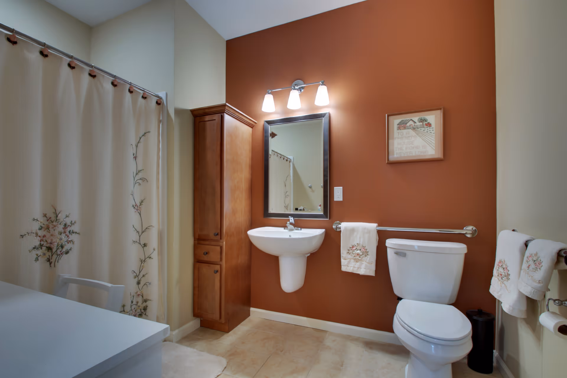 A bathroom with a white toilet, a wall-mounted white sink, a wooden cabinet, and a mirror with three lights above it. The walls are painted in a combination of beige and burnt orange. There are floral-patterned towels hanging on metal bars and a shower curtain with a floral design.