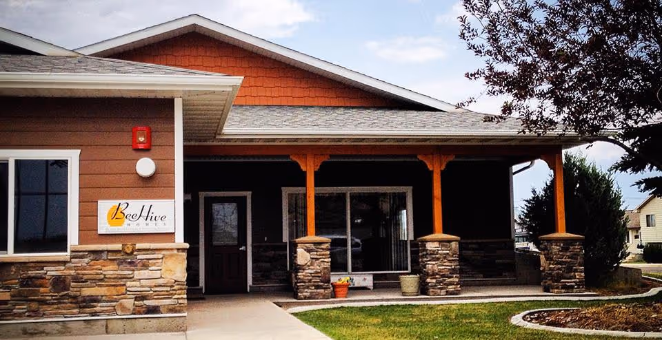 Front entrance of BeeHive Homes with a covered porch, stone columns, wooden posts, and a small lawn.