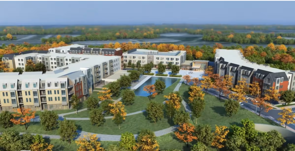 Aerial view of The Baldwin Senior Living facility showing multiple connected buildings surrounded by landscaped green areas with trees and walking paths. There is a central water feature and outdoor seating areas, with a backdrop of forested land and open fields.
