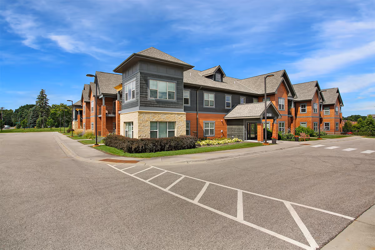 Exterior view of a two-story senior living facility building with a mix of stone, wood, and siding finishes under a blue sky with some clouds. The building has multiple windows, a covered entrance, and is surrounded by a paved parking area and landscaped greenery.