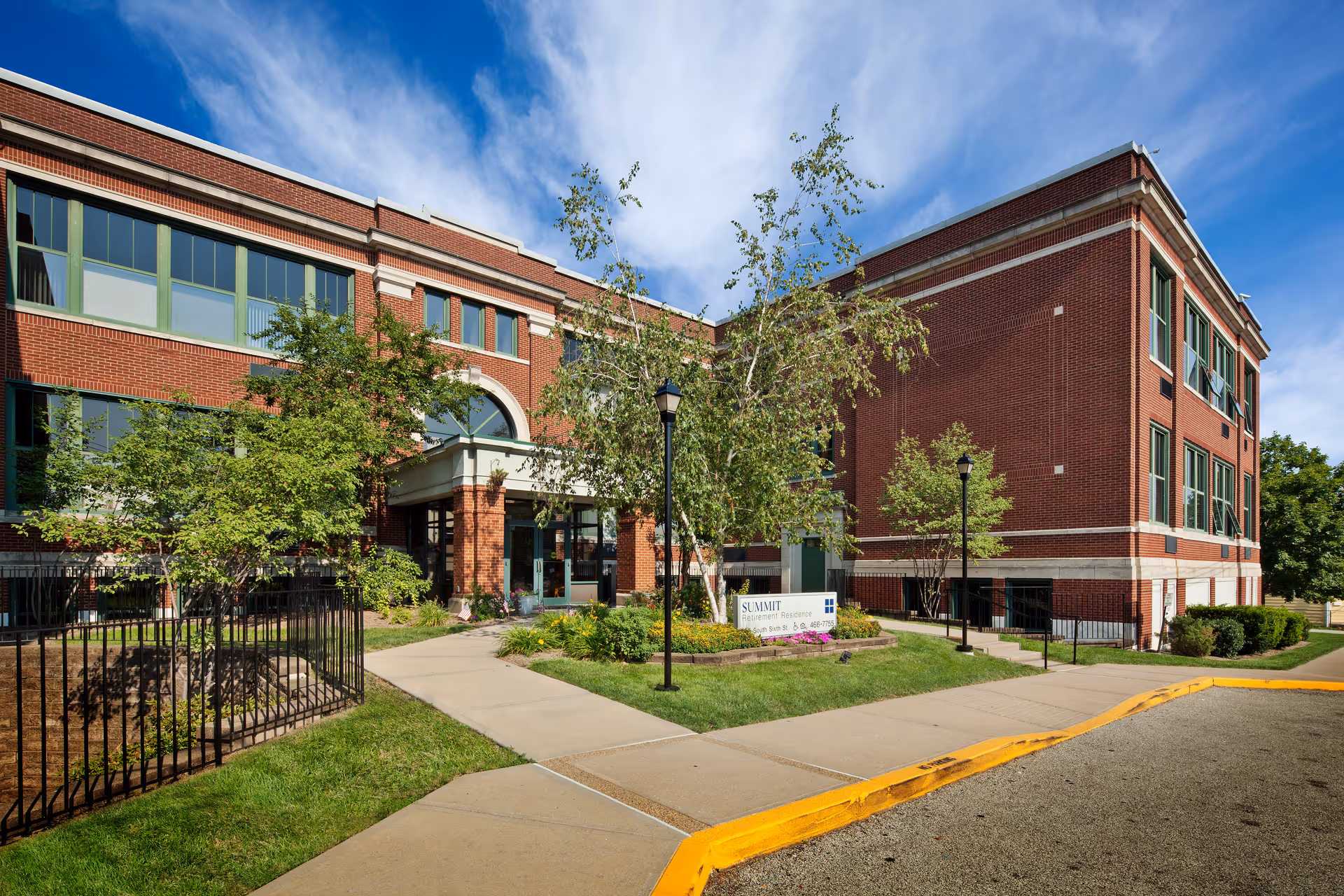 Exterior view of Summit Retirement Residence, a red brick building with large windows and a landscaped entrance featuring trees, shrubs, and a sidewalk leading to the main door under a clear blue sky.