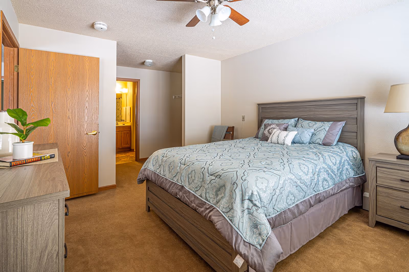 A bright furnished bedroom with a patterned bed, wooden nightstand and dresser, ceiling fan, and an open door showing a bathroom.