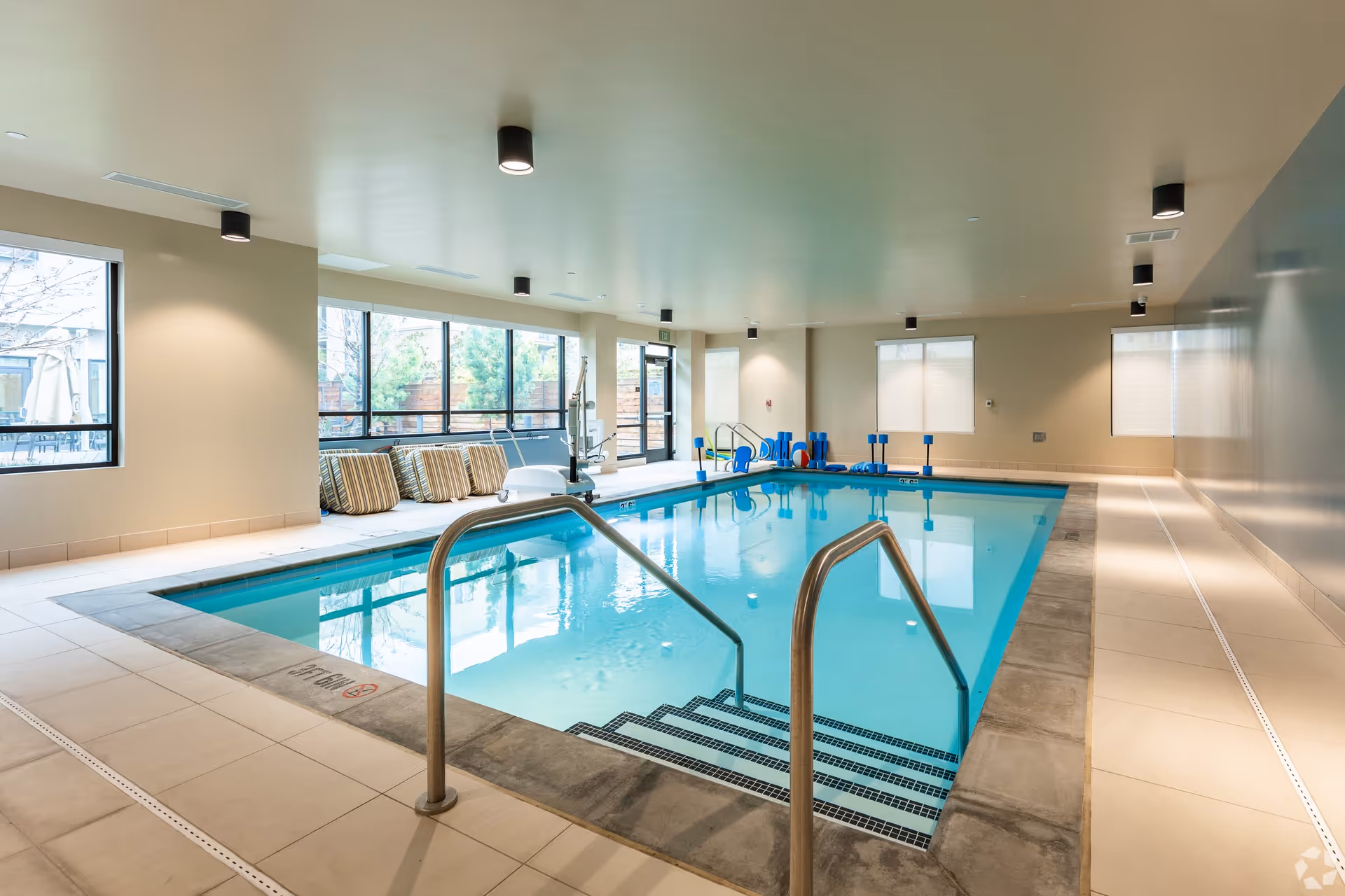 Indoor swimming pool with metal handrails and steps leading into the water, surrounded by beige tiled flooring and walls. Large windows on one side let in natural light, with some pool equipment and striped cushions placed near the windows.