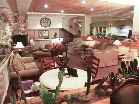 A cozy senior living facility common area with multiple beige sofas and armchairs arranged around coffee tables. There is a stone fireplace on the left side and a round table with chairs in the foreground, decorated with a small eagle statue and various plants. The room has warm lighting with lamps on tables and recessed ceiling lights. A staircase and hallway are visible in the background.