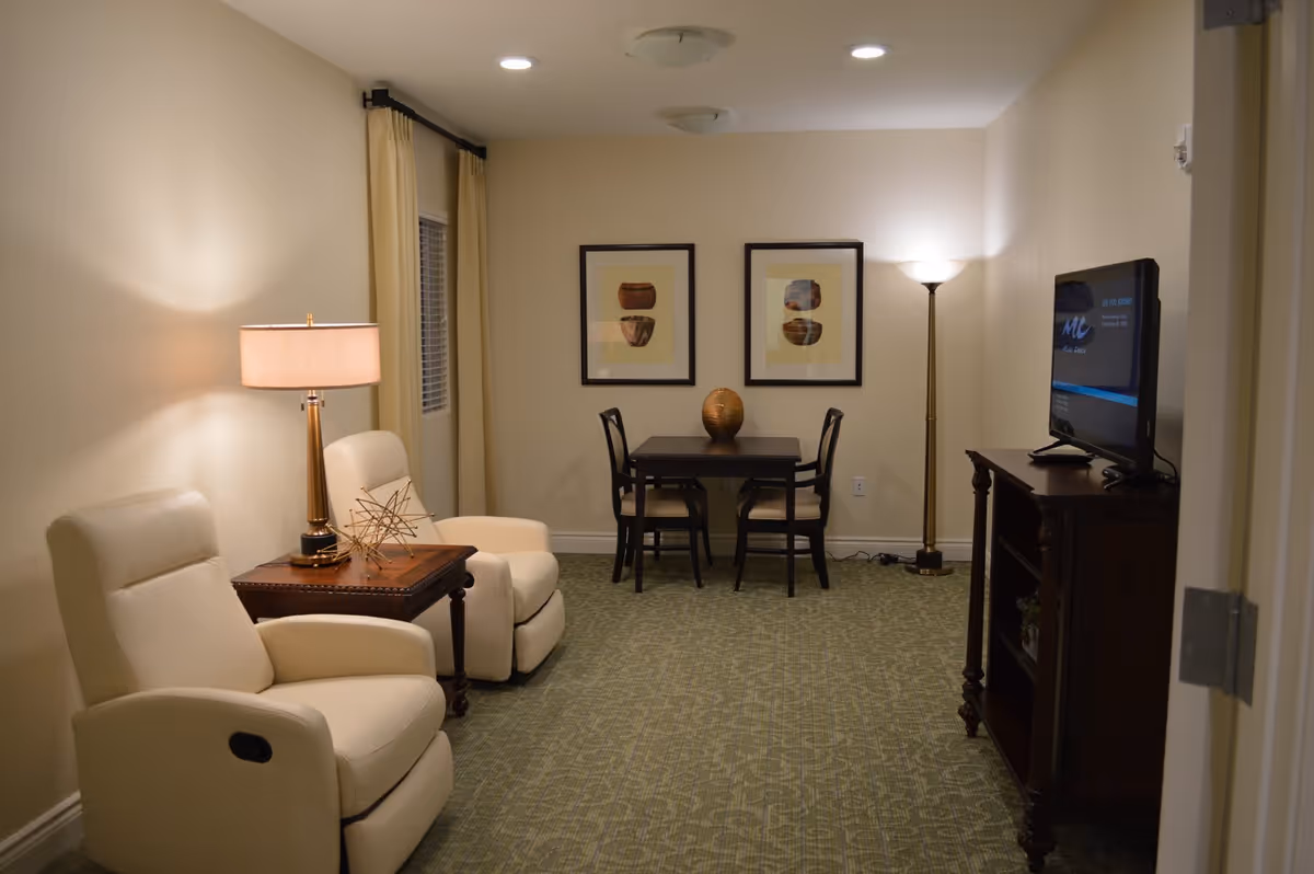 A cozy living room area with two cream-colored recliner chairs separated by a wooden side table with a lamp and decorative object. In the background, there is a small dark wooden table with two chairs and a decorative vase. Two framed artworks hang on the wall above the table. A floor lamp stands in the corner next to a wooden TV stand with a flat-screen television. The room has beige walls and green patterned carpet.