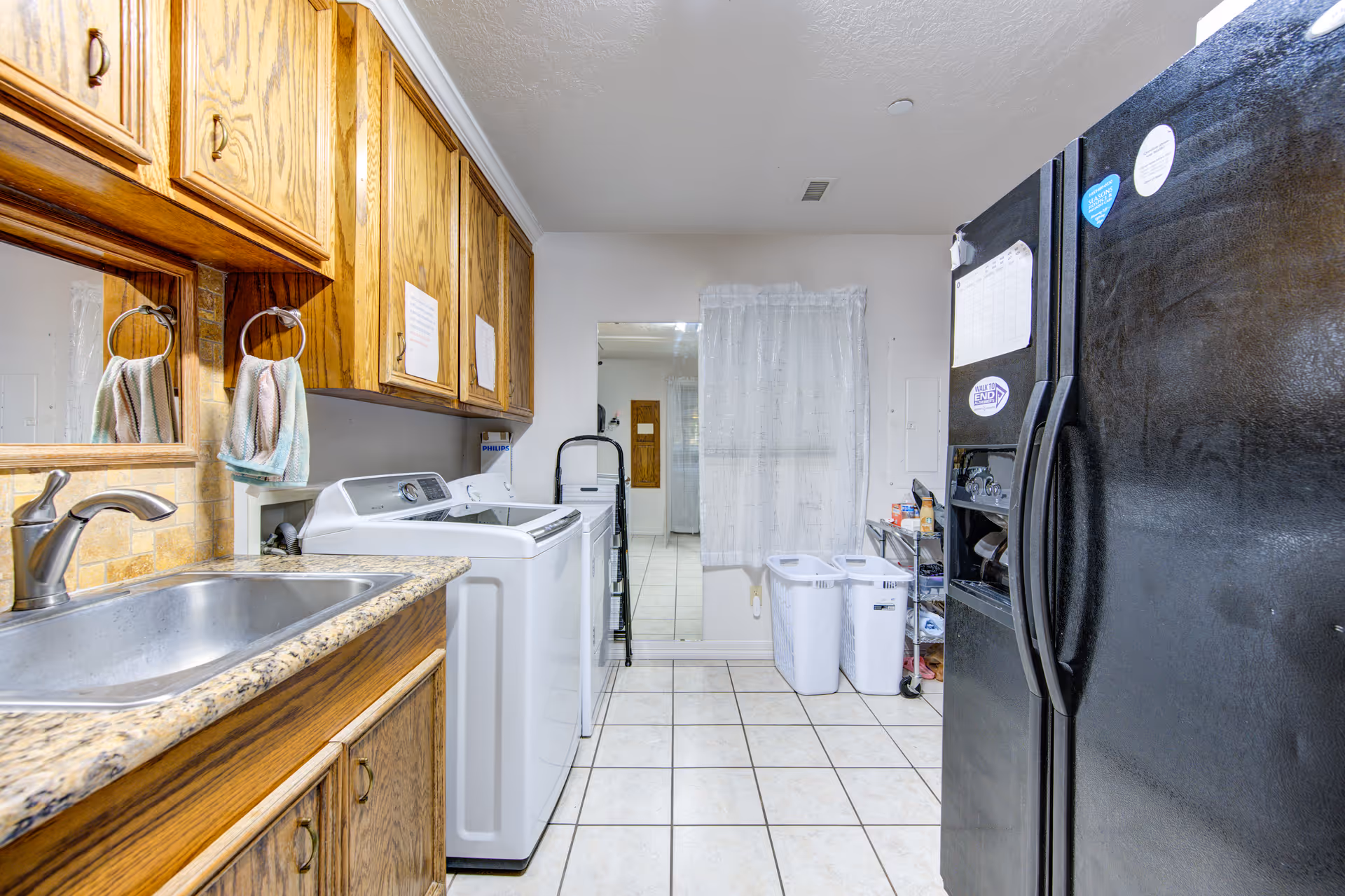 Laundry room with a washing machine and dryer side by side, wooden cabinets above the machines, a stainless steel sink with a granite countertop, a black refrigerator on the right, two white laundry baskets, and a window with a sheer white curtain.