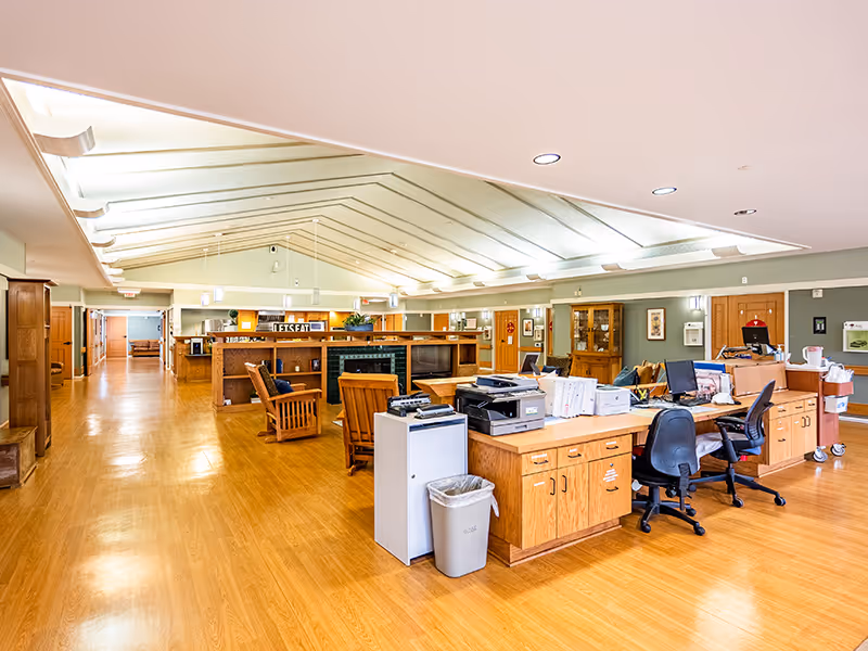 A spacious interior area with wooden flooring and a high ceiling with skylights. The room features a reception or office desk area with office chairs, printers, and paperwork. There are wooden chairs and tables arranged in the background, along with cabinets and framed pictures on the walls. The space is well-lit with ceiling lights and natural light from the skylights.