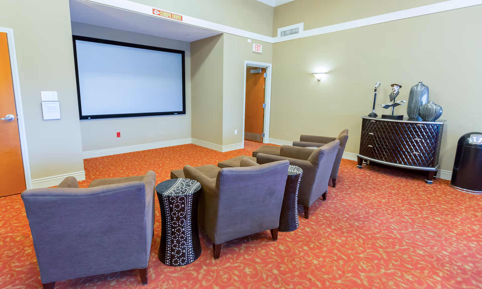 Small screening room with a blank projector screen, a row of upholstered chairs, side tables, and a decorative cabinet against the wall.