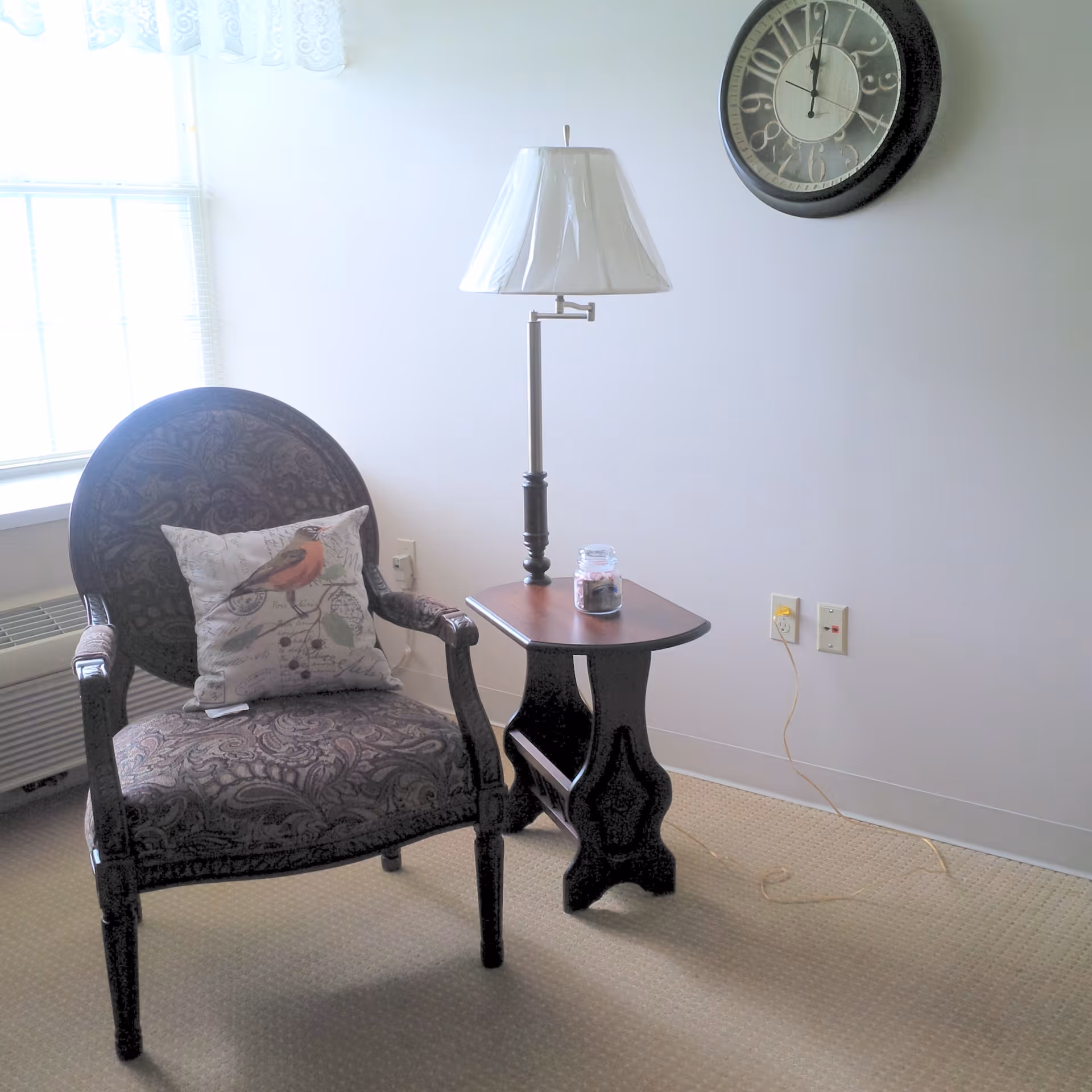Upholstered armchair with a decorative bird pillow next to a small side table holding a lamp and jar, under a wall clock in a bright room.