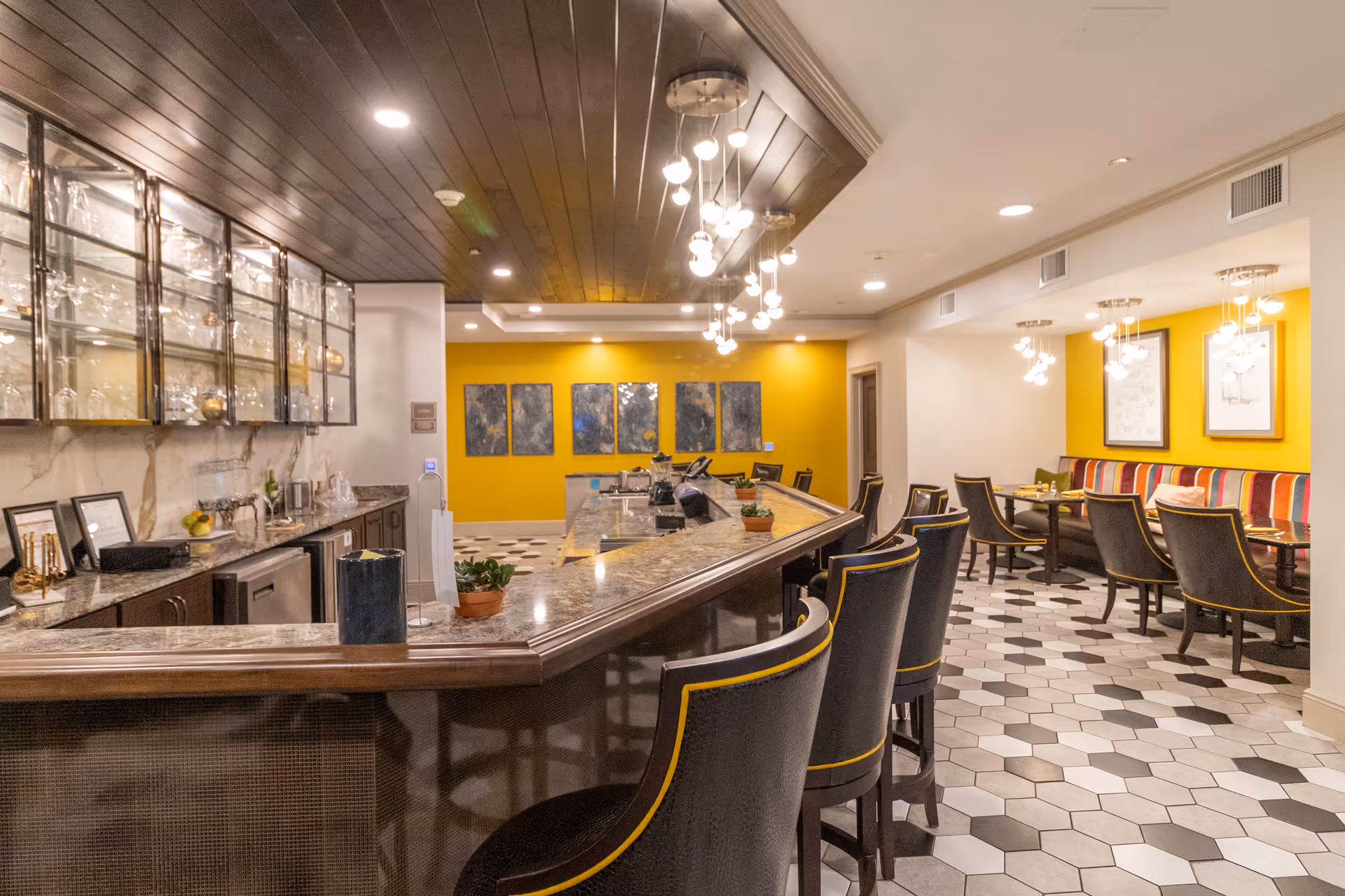 Well-lit modern dining and bar area with a curved marble countertop, high-backed stools, yellow accent walls, and hexagonal tile flooring.