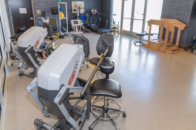 A bright interior therapy/exercise room with seated exercise machines, rolling stools, and additional equipment near glass doors.