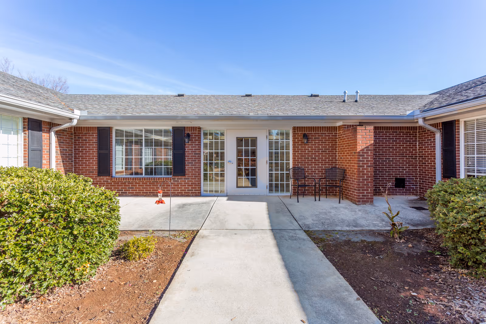 Exterior view of a single-story brick building with a central glass door flanked by large windows with black shutters. There are two chairs and a small table on the right side of the entrance. The building is surrounded by trimmed bushes and a concrete pathway leading to the door under a clear blue sky.