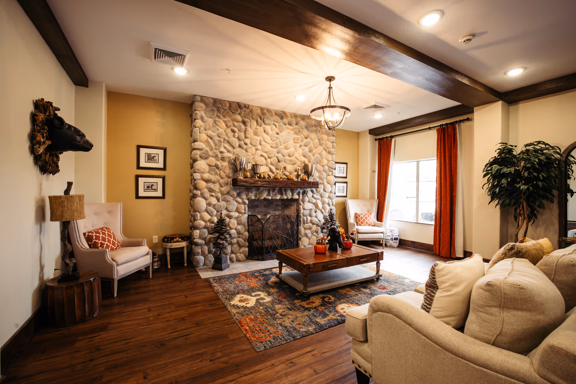 A cozy living room with a stone fireplace as the focal point, flanked by two beige armchairs with orange patterned pillows. A wooden coffee table sits on a colorful rug in front of a beige sofa with multiple cushions. The room features wooden flooring, a large window with orange curtains, a mounted animal head on the wall, a floor lamp, and a potted plant in the corner.