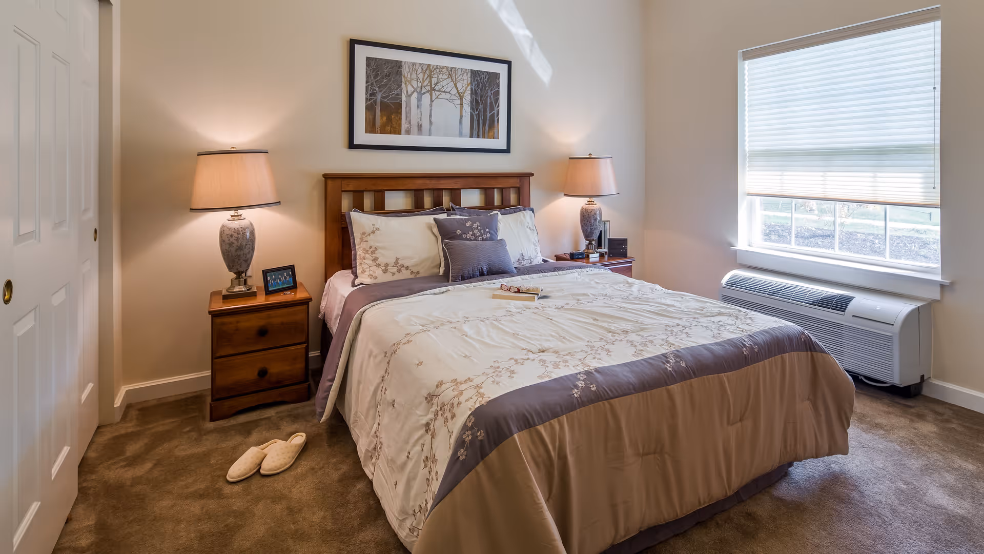 A neatly made bed with floral-patterned bedding in a bedroom. There are two wooden nightstands on either side of the bed, each with a lamp. A framed picture of trees hangs above the bed. A window with blinds is on the right wall, and a pair of slippers is on the carpeted floor near the left nightstand.