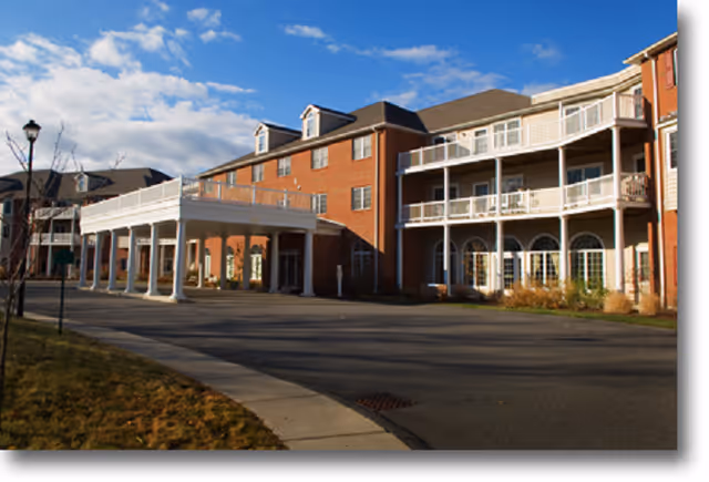 Exterior view of a multi-story brick building with white balconies and a covered entrance, under a partly cloudy blue sky.