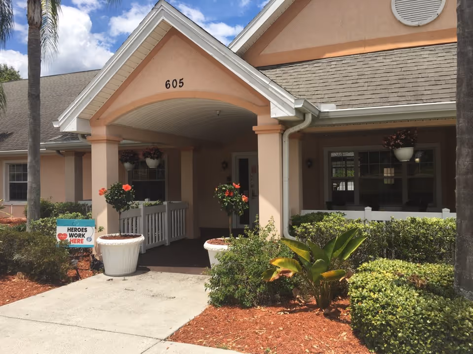 Front entrance of a peach-colored building with the number 605 above the covered porch. There are two large white planters with flowering plants on either side of the walkway leading to the door. Green shrubs and palm trees surround the entrance. A sign near the walkway reads 'HEROES WORK HERE'.