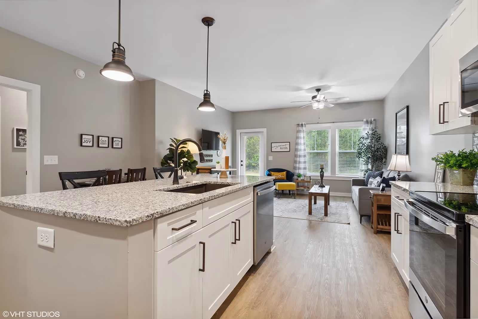 Modern open-concept kitchen and living room area with a large granite countertop island featuring a sink and dishwasher. The kitchen has white cabinets and stainless steel appliances. The living room has a gray sofa, a yellow armchair, a coffee table, and large windows with curtains letting in natural light. Pendant lights hang over the island, and a ceiling fan is visible in the living room.