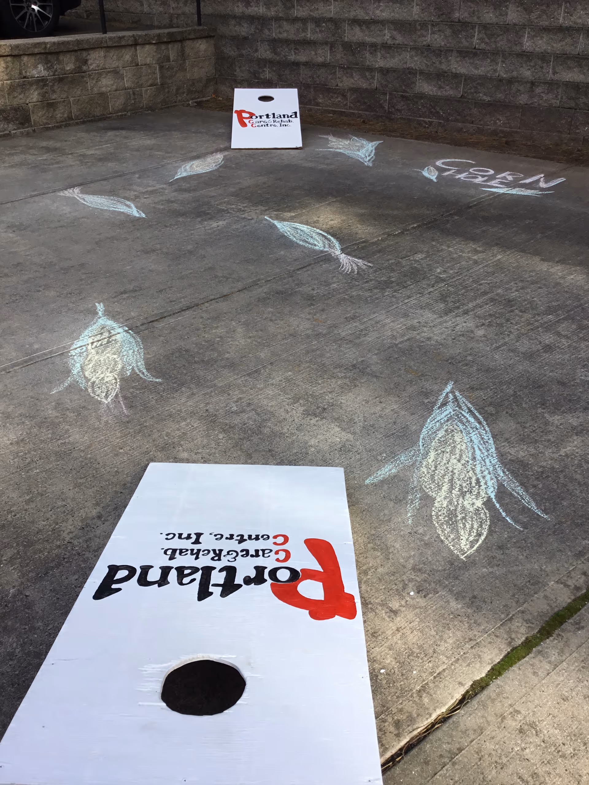Concrete outdoor area with two cornhole boards labeled "Portland Care & Rehab Centre, Inc." and chalk drawings of ears of corn on the ground.
