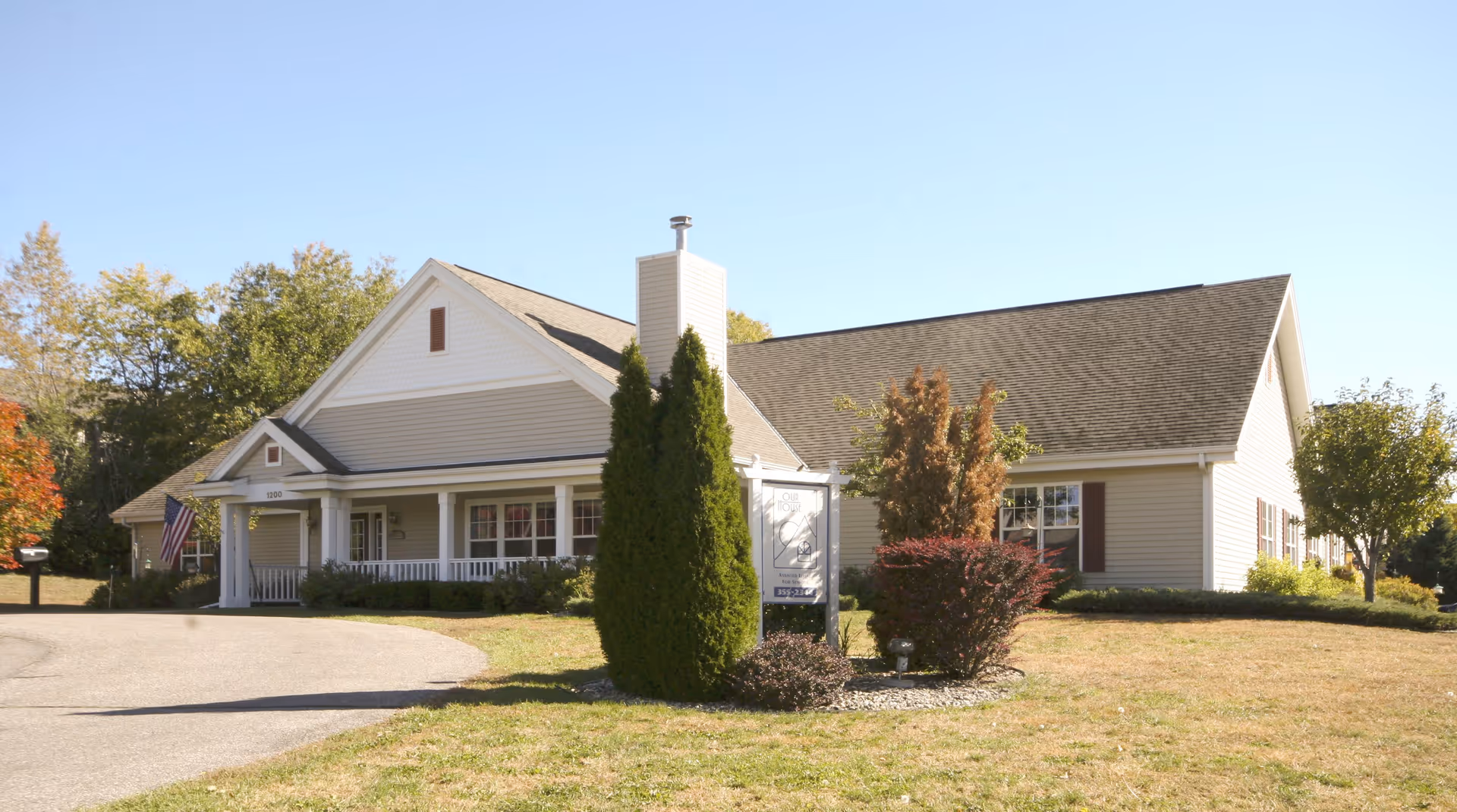 Single-story beige assisted living building with a covered front porch, driveway, and landscaped lawn under a clear sky.