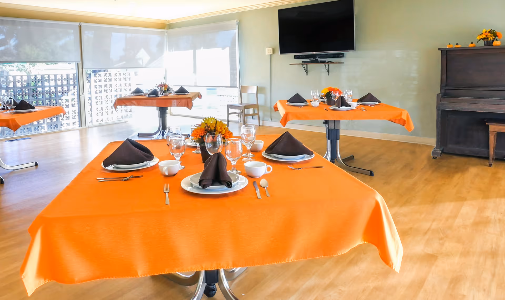 A bright dining room with several tables covered in orange tablecloths, each set with plates, glasses, cups, silverware, and folded dark napkins. The room has large windows with white blinds, a wall-mounted TV, a wooden piano, and wooden flooring.