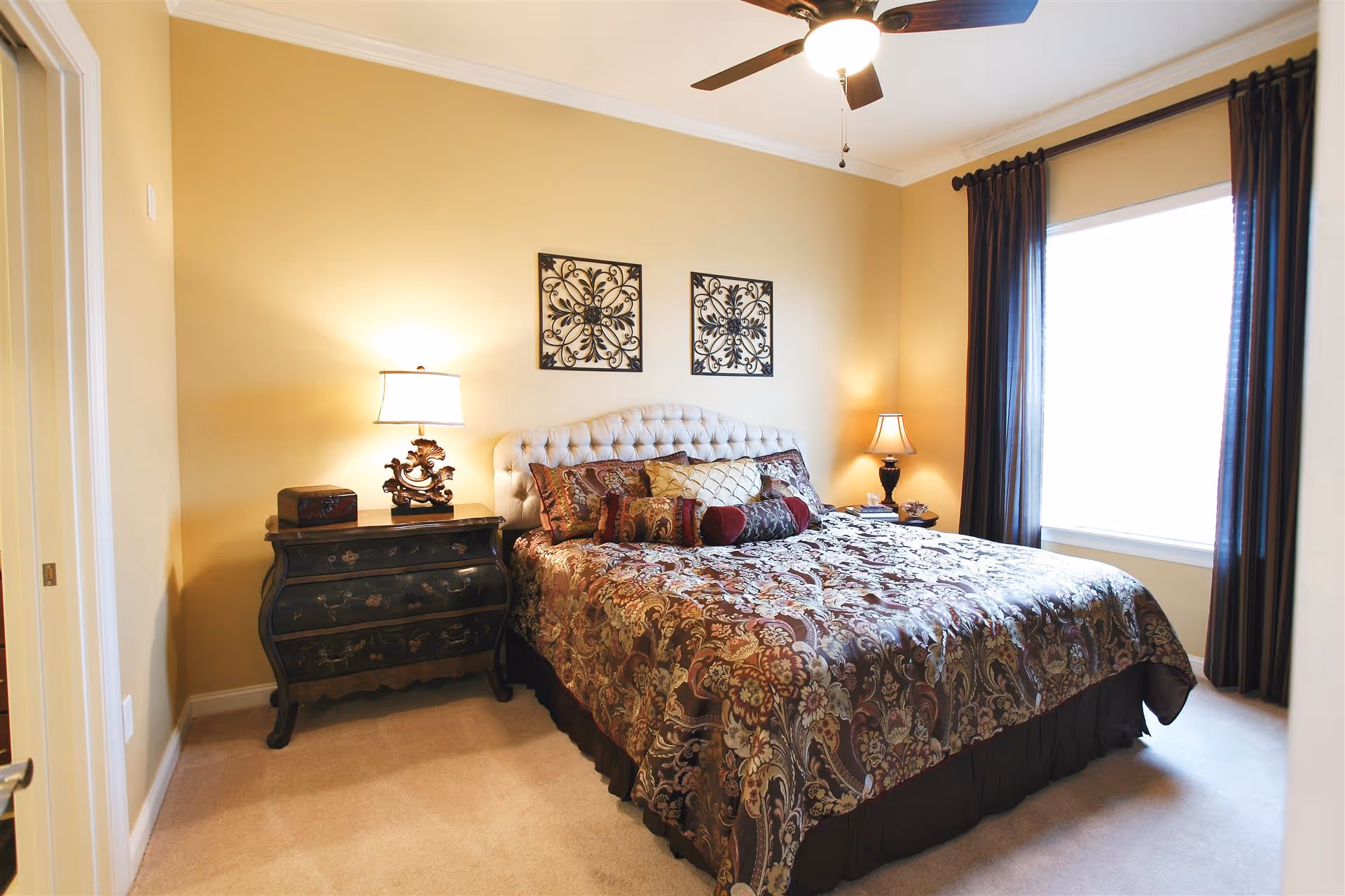 A cozy bedroom with a large bed covered in a patterned bedspread and multiple decorative pillows. There are two bedside tables, each with a lamp, and two decorative wall hangings above the tufted headboard. A ceiling fan with a light is mounted on the ceiling, and a large window with dark curtains allows natural light into the room.