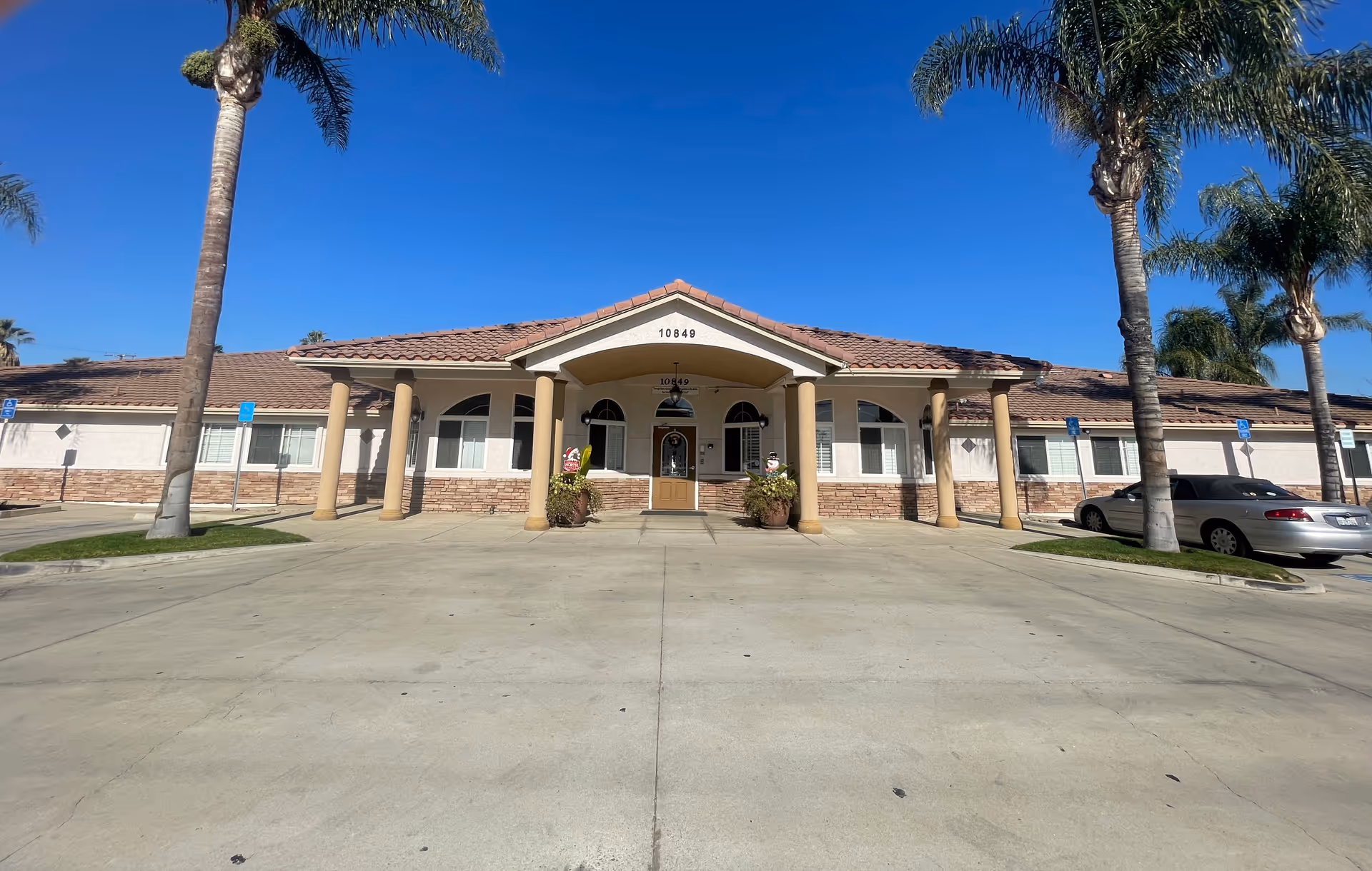 Front exterior view of a single-story building with a tiled roof and beige columns supporting a covered entrance. The building has several windows with white shutters and is flanked by palm trees. There are a few cars parked on the right side and handicap parking signs visible. The sky is clear and blue.
