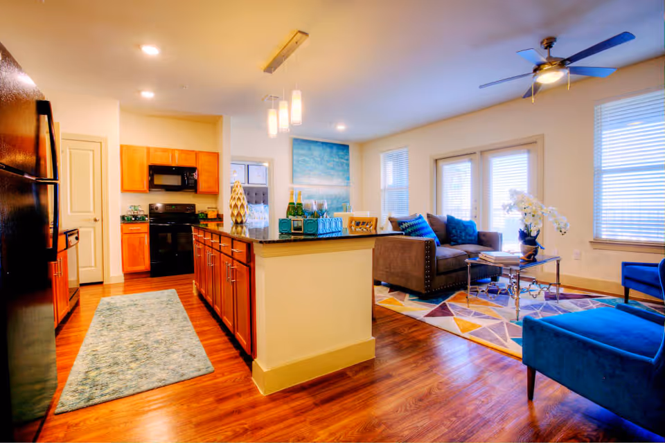 Bright and modern open-concept living space featuring a kitchen with wooden cabinets, black appliances, and a central island with decorative items. Adjacent to the kitchen is a cozy living room area with a gray sofa, blue armchair, colorful geometric rug, glass coffee table, and large windows with blinds allowing natural light to fill the room.