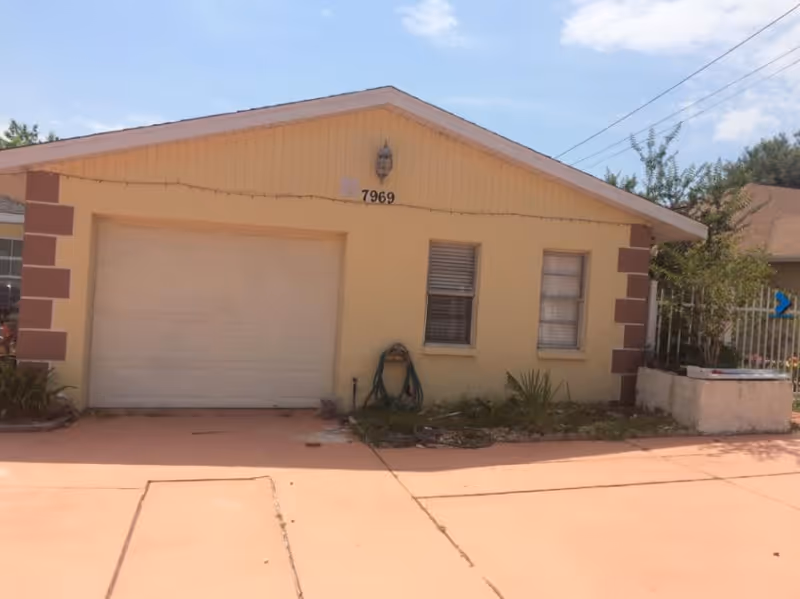 Exterior view of a single-story building with a light yellow facade and a white garage door. The building has two windows with blinds, a garden hose coiled on the wall, and some plants in front. The address number 7969 is displayed above the garage door. The sky is clear with a few clouds.