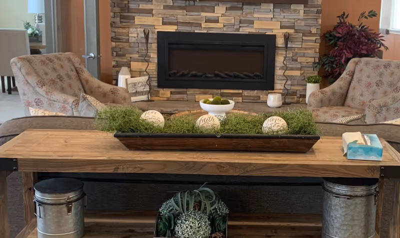 A cozy living room area with two floral-patterned armchairs facing a stone fireplace. In front of the chairs is a wooden console table decorated with a long tray filled with green moss and three decorative balls labeled 'Love', 'Faith', and 'Hope'. There is also a small bowl with green decorative balls and a box of tissues on the table. The room has warm lighting and a plant in the corner near a window.