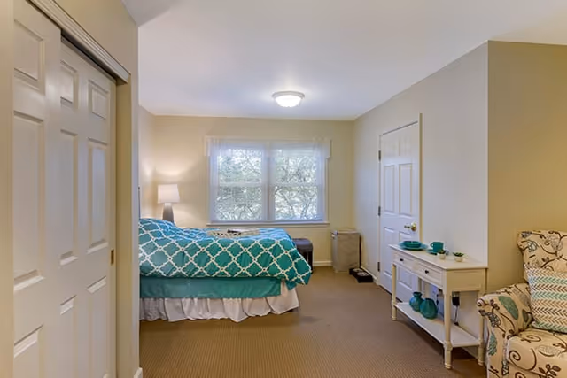A cozy bedroom with a bed covered in a teal and white patterned comforter, a window with white curtains letting in natural light, a small white console table with decorative items, and a patterned armchair with a teal pillow. The room has beige walls and carpet, with a closet door on the left and a closed door on the right.