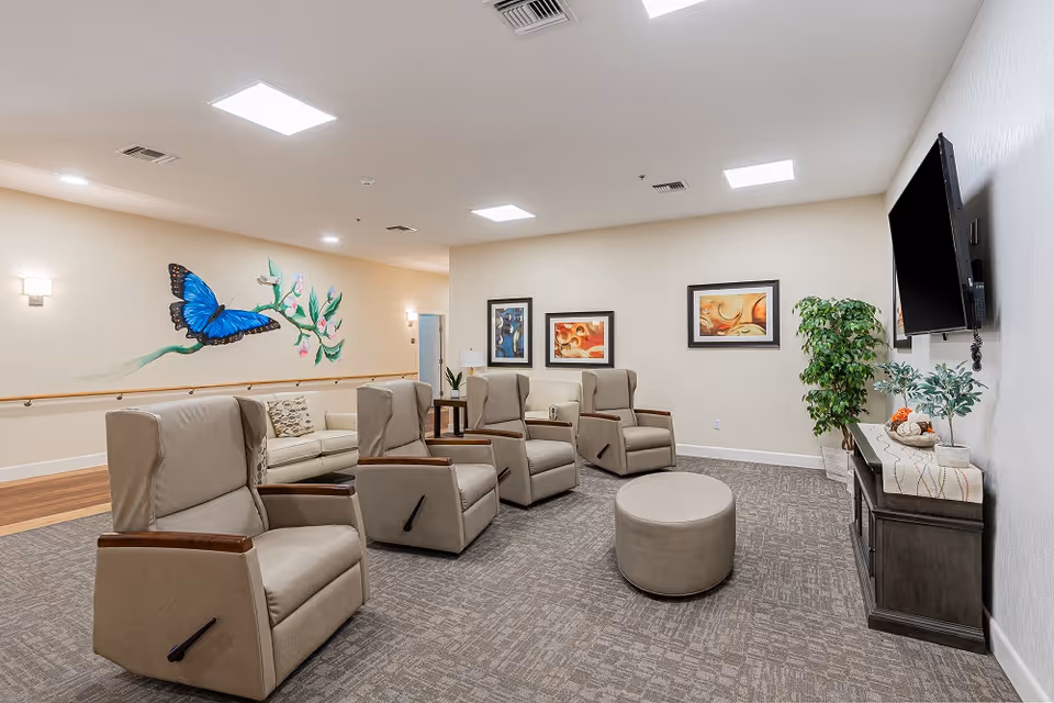 A well-lit living room area in an assisted living facility featuring four beige recliner chairs arranged in a semi-circle around a round ottoman. There is a beige sofa against the wall with a colorful butterfly mural above it. The walls are decorated with framed abstract art, and a flat-screen TV is mounted on the right wall above a dark wooden cabinet with plants and decorative items. The room has carpeted flooring and ceiling lights.