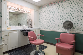 A small salon or grooming room with two pink salon chairs, a black hair washing sink, a large mirror with lights above it, white cabinets below the sink, and floral wallpaper on the walls.