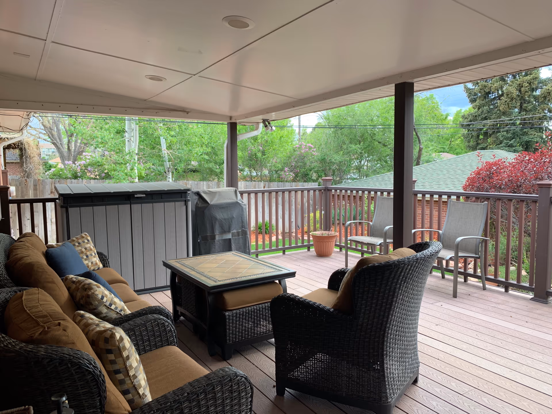 Covered outdoor patio area with wicker furniture including a sofa and chairs with cushions, a square coffee table, two metal chairs, a storage container, a covered grill, and potted plants. The patio overlooks a fenced backyard with green trees and shrubs.