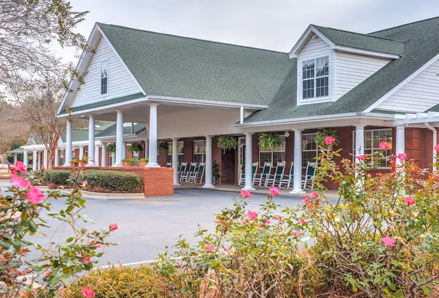 Exterior view of a senior living facility with a covered entrance supported by white columns, green rocking chairs on the porch, hanging plants, and blooming pink flowers in the foreground.