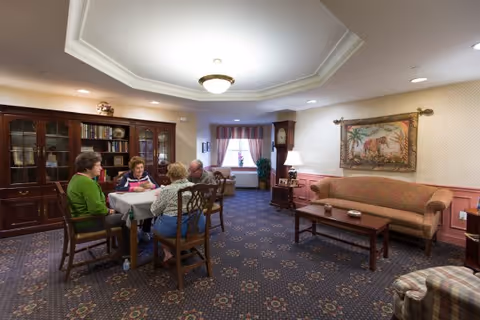 Four seniors seated around a table playing cards in a furnished community living room with a sofa and bookshelves.