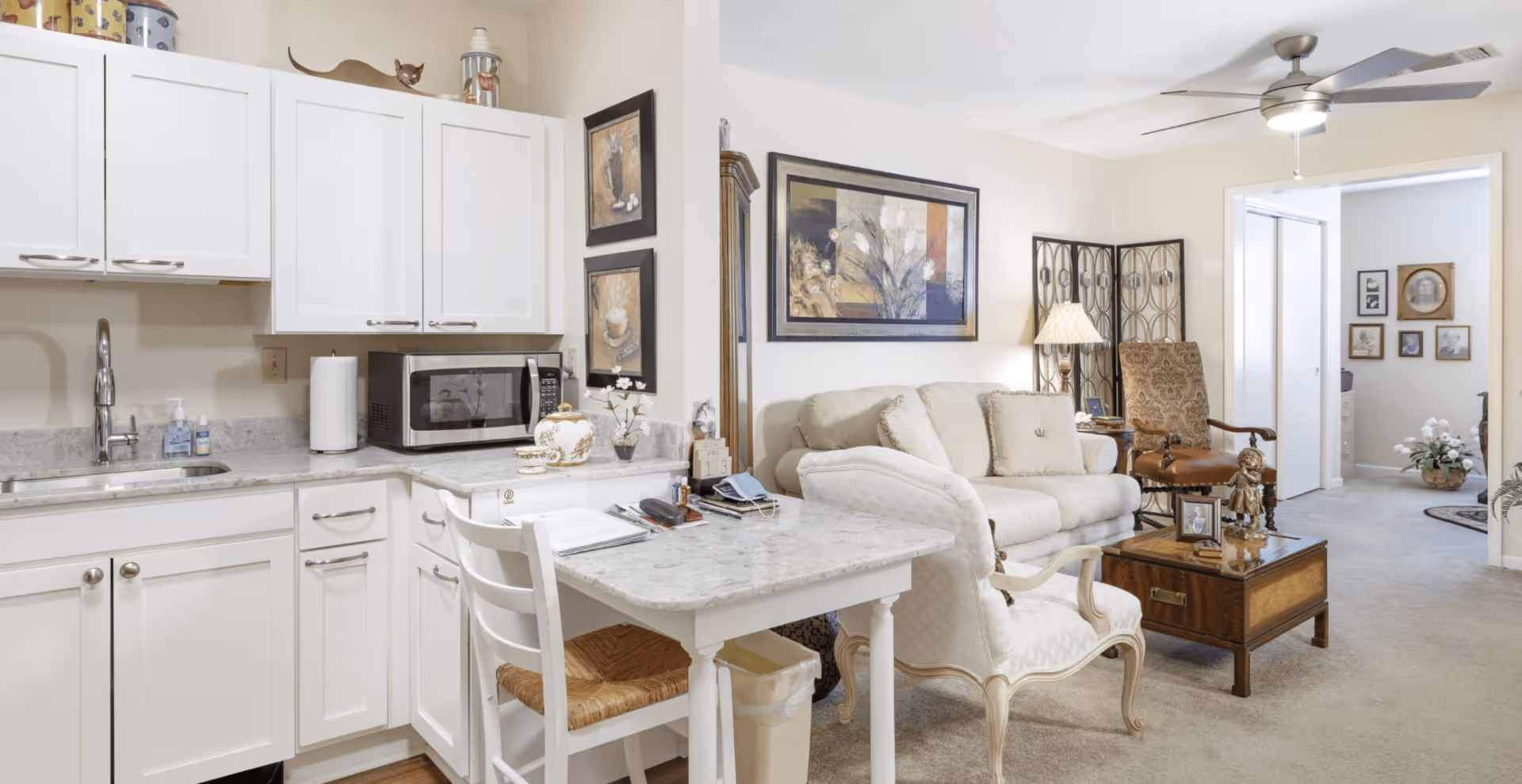 A cozy living space featuring a small kitchen area with white cabinets, a marble countertop, a microwave, and a sink. Adjacent to the kitchen is a small table with a white chair. The living room area includes a beige sofa, an armchair, a wooden coffee table with decorative items, a floor lamp, and framed artwork on the walls. A ceiling fan with a light is mounted on the ceiling. In the background, another room is visible with framed pictures on the wall and a floral arrangement on the floor.