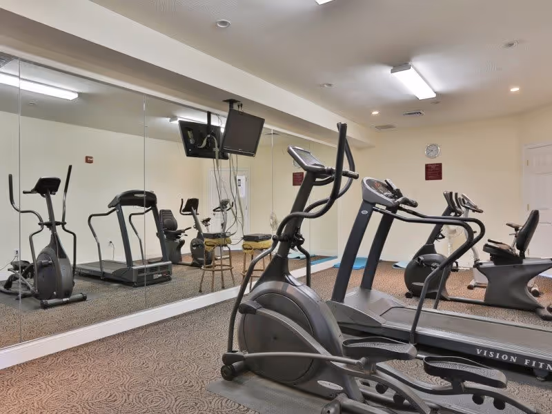 Interior view of a fitness room at Merrimack Meadows with exercise equipment including treadmills, stationary bikes, and elliptical machines. The room has a large wall mirror, beige walls, and carpeted floor. A wall clock and a mounted TV are also visible.