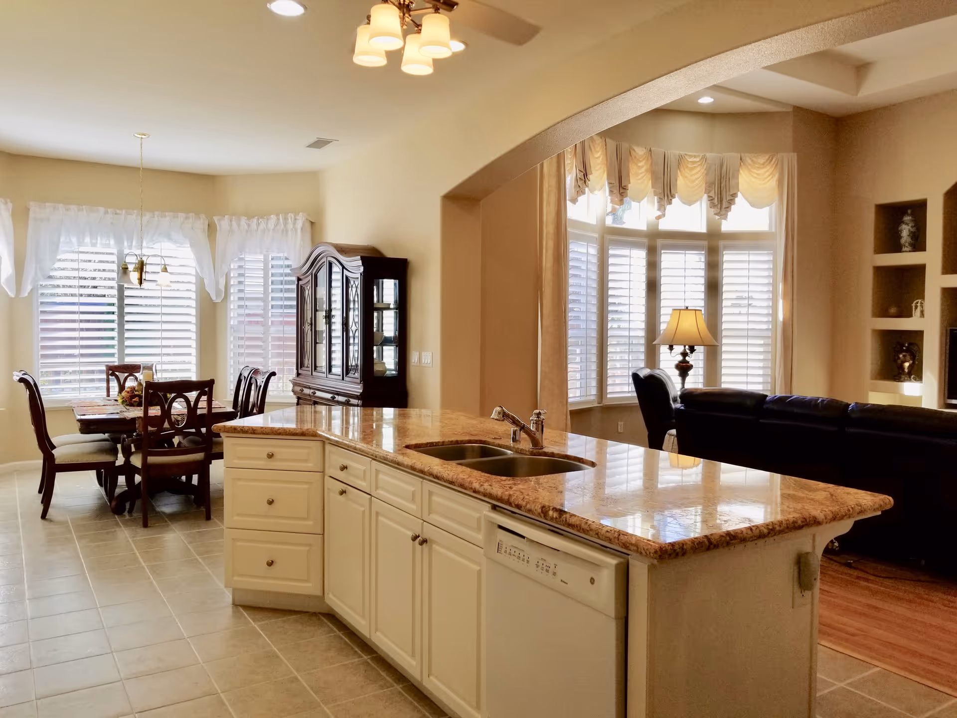 Interior view of a senior living facility showing a kitchen island with a double sink and dishwasher, adjacent to a dining area with a wooden dining table and chairs, and a living room with a black leather sofa and a lamp near large windows with white blinds and curtains.